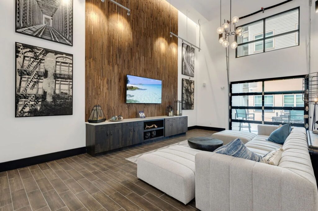 Modern living room with high ceiling, wood-paneled accent wall, sectional sofa, wall-mounted TV, and large windows. Artworks and pendant lights add decorative touches.