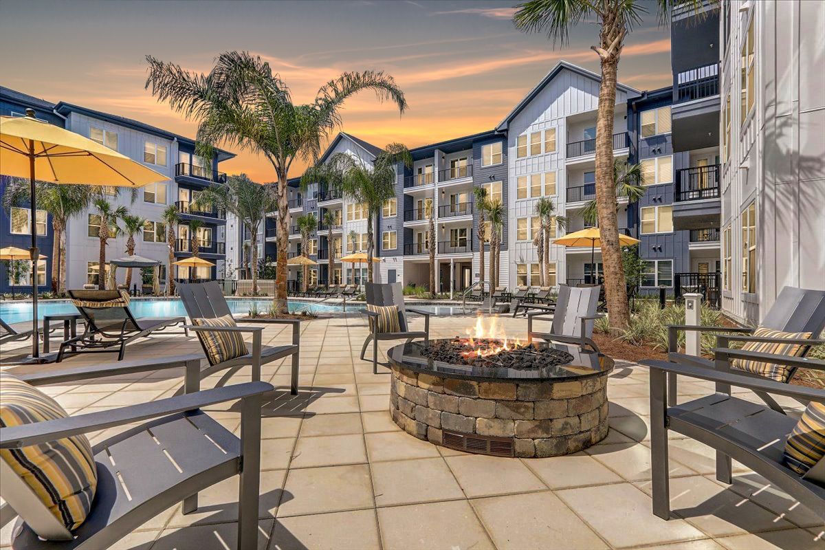 Outdoor seating area with a fire pit and chairs, surrounded by apartment buildings and palm trees, at sunset.